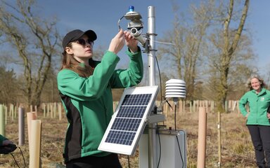A woman installs a sensor onto a tripod containing a solar panel, equipment box and various cables.