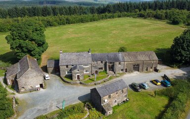 A drone shot of a farmhouse-style building with field and forest behind