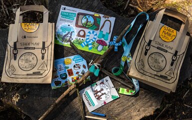 Stick Man themed postcard, lanyard, stickers and leaflet laid out on a log slice.
