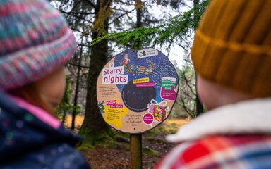 Two children in winter coats and hats looking at a Stick Man panel in the forest.