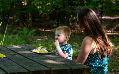 A woman and young boy sitting on a wooden picnic bench in a forest. The child is eating crisps.