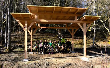 group of people sat under a large timber shelter in a forest
