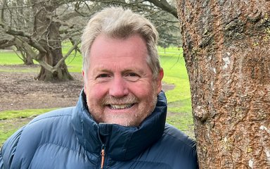 A mann with grey hair and a short grey beard leans against a tree. he smiles at the camera.