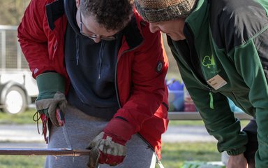 A young man dressed in a red coat stares down at a long piece of wood that he is sawing to fit to size. A gentleman in a green coat watches over to support him.