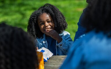 A young girl smiling as she eats crisps at a picnic bench.