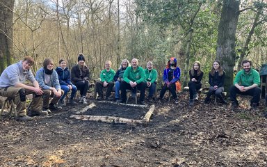 A group of teenagers and adults sit round a square unlit fire pit in the middle of a forest setting, smiling at the camera.