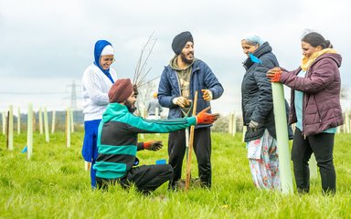 A group of five adults at a tree planting event, one kneeling, one with a spade and one holding a tree sapling in protective material.