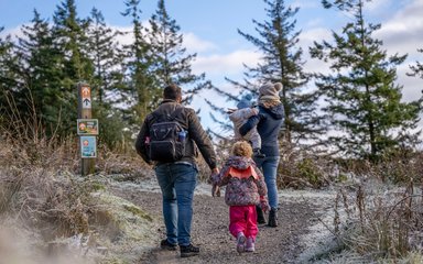 With their backs to the camera, a family of two adults and two children walking on a forest path edged with frost. One of the children is wearing orange wings.