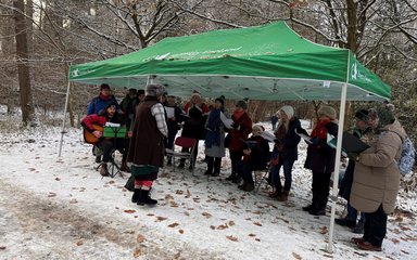 A group of people wearing hats and coats singing under a gazebo in the forest, with a dusting of snow on the ground