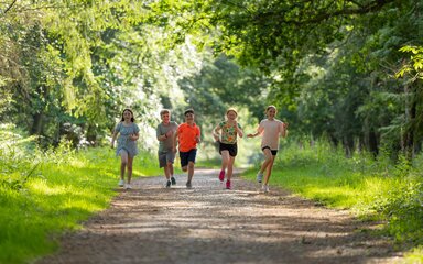 A summer image of five children running on a forest trail surrounded by trees in with dappled sunlight.