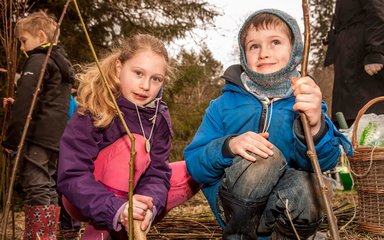 A young boy and girl face the camera, holding sticks as part of a bushcraft activity