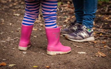 A close-up of two pairs of children's legs in the forest, one wearing pink wellington boots and the other blue trainers.
