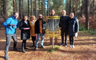 Seven people, some holding frisbees, smile at the camera next to a disc golf net in the forest