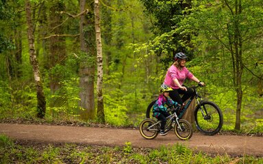 A woman and child cycling along a forest path, surrounded by green trees.