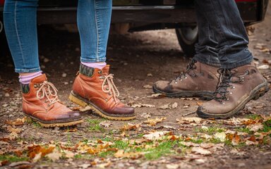 Close-up of two people's walking boots stood on forest ground.