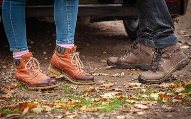 Close-up of two people's walking boots stood on forest ground.