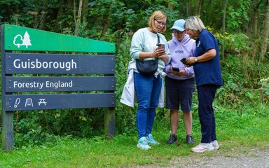 Three people stood together by a Forestry England sign for Guisborough Forest.