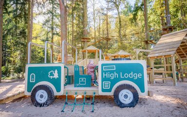 Young girl sat in a wooden truck play structure in a sandy play area.