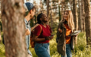 Three young people walk and talk through a forest landscape. 