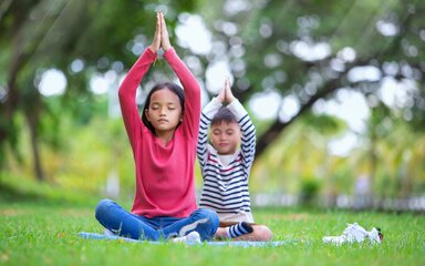 2 children sit crossed legged in a woodland environment with eyes closed and hands over their heads