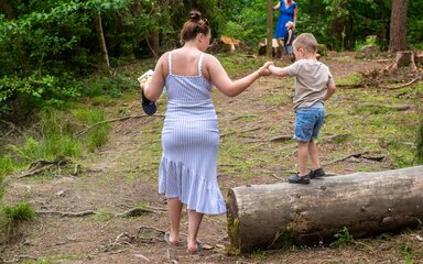 A woman holds the hand of a young boy climbing on a fallen log in the forest.