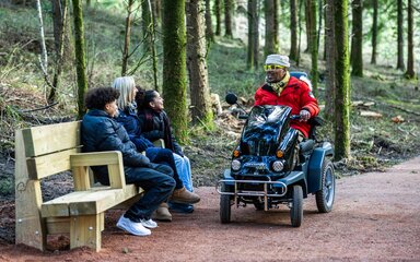 A man on an all-terrain mobility scooter talks to a woman and two children sitting on a bench in the forest.