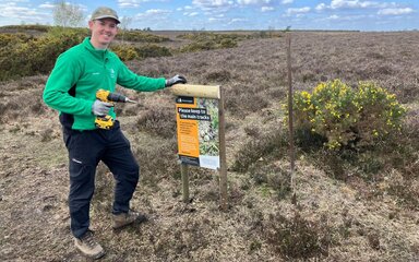 A man wearing Forestry England uniform, smiling as he holds a drill next to a wooden signpost outdoors.