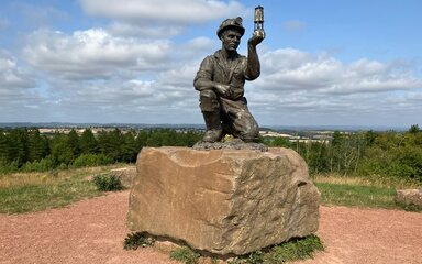 Bronze miner sculpture holding a lamp on a stone plinth at Silverhill Wood, with panoramic countryside views behind.