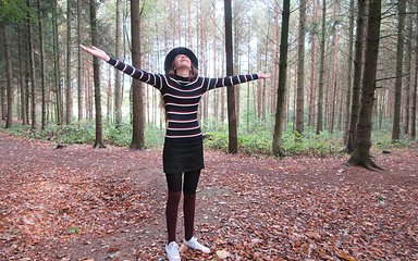 A teenage girl wearing a stripy long sleeved top, a short black skirt with black tights and white trainers hods her arms out wide joyfully in front of a lot of leafless brown thin trees. The ground is brown and covered in leaves.