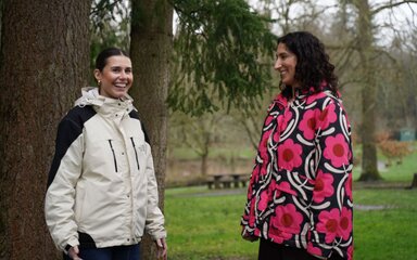 Two women smiling at each other, standing under a tree in the forest.