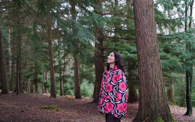 A woman in a colourful coat smiles as she looks up at trees in the forest.