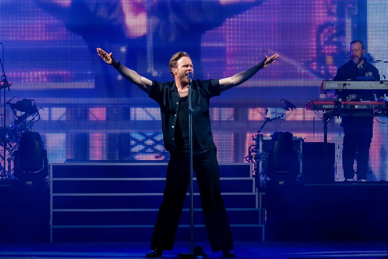 Male singer Olly Murs standing on stage in front of a microphone, with his arms spread.