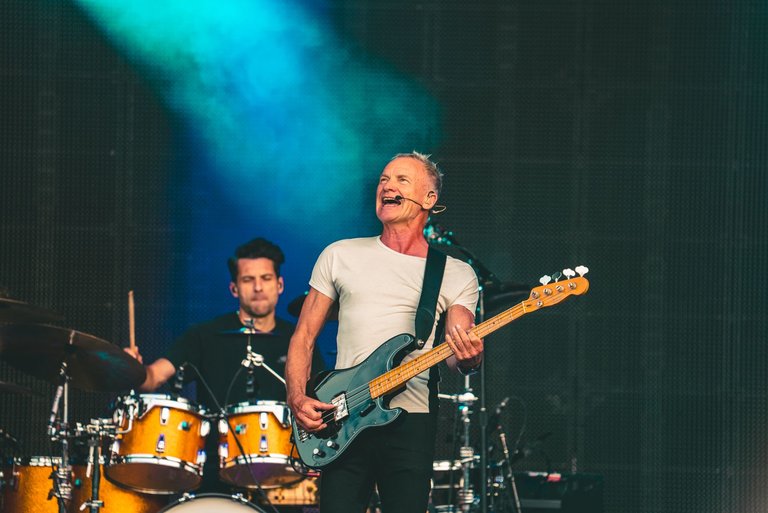 Male singer Sting holding on electric guitar on stage, with a male drummer at a drum kit sat behind.