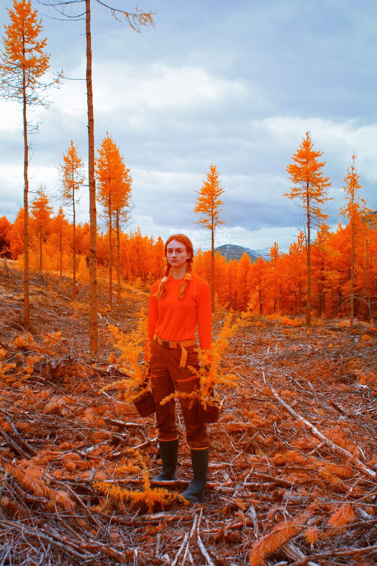A red-haired woman wearing orange standing near trees with orange foliage.