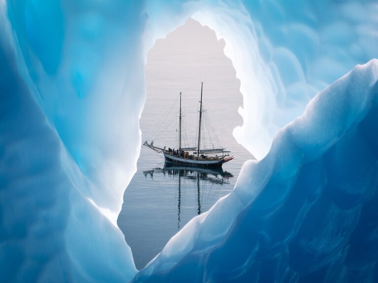 A tall ship with sails down, seen through a gap among ice bergs.