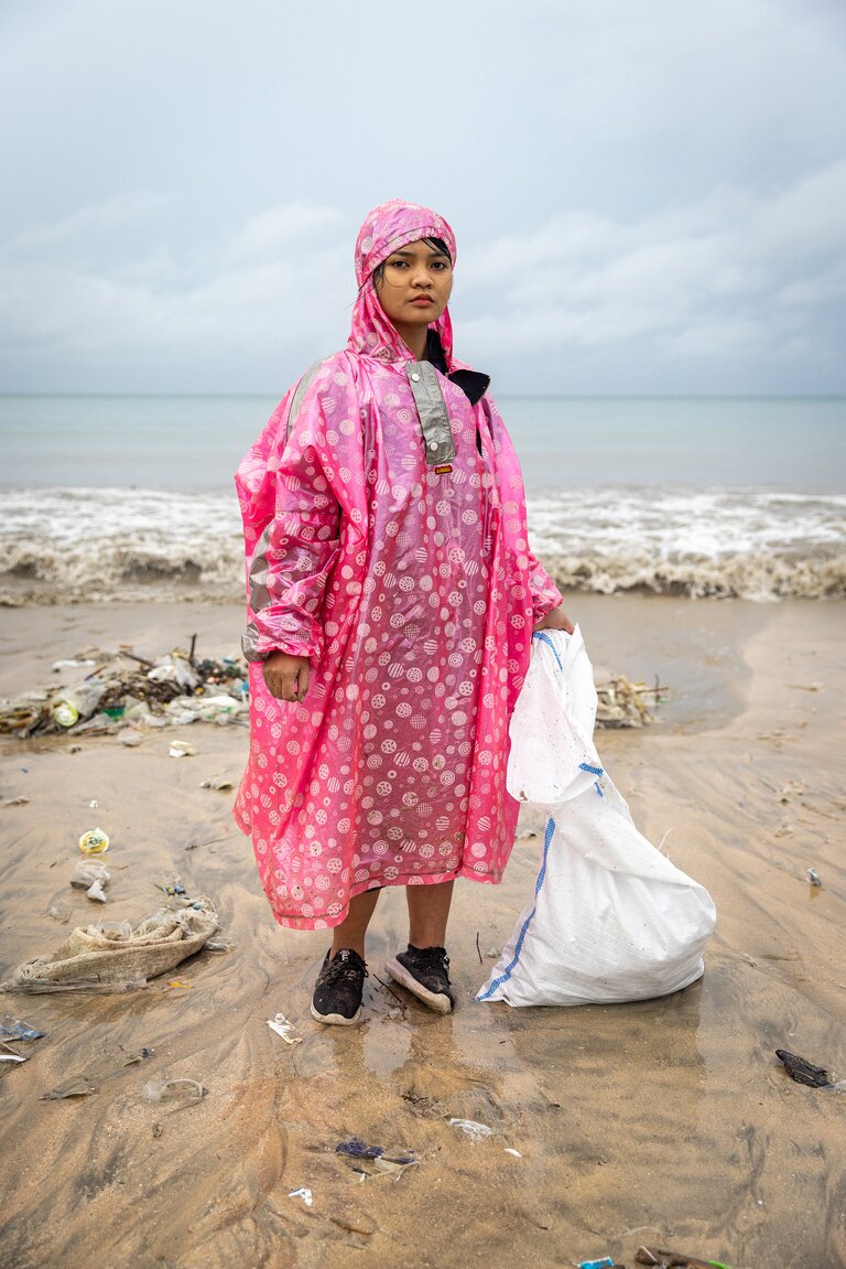 A woman wearing a pink waterproof poncho standing on a littered sea shore, holding a plastic bag.