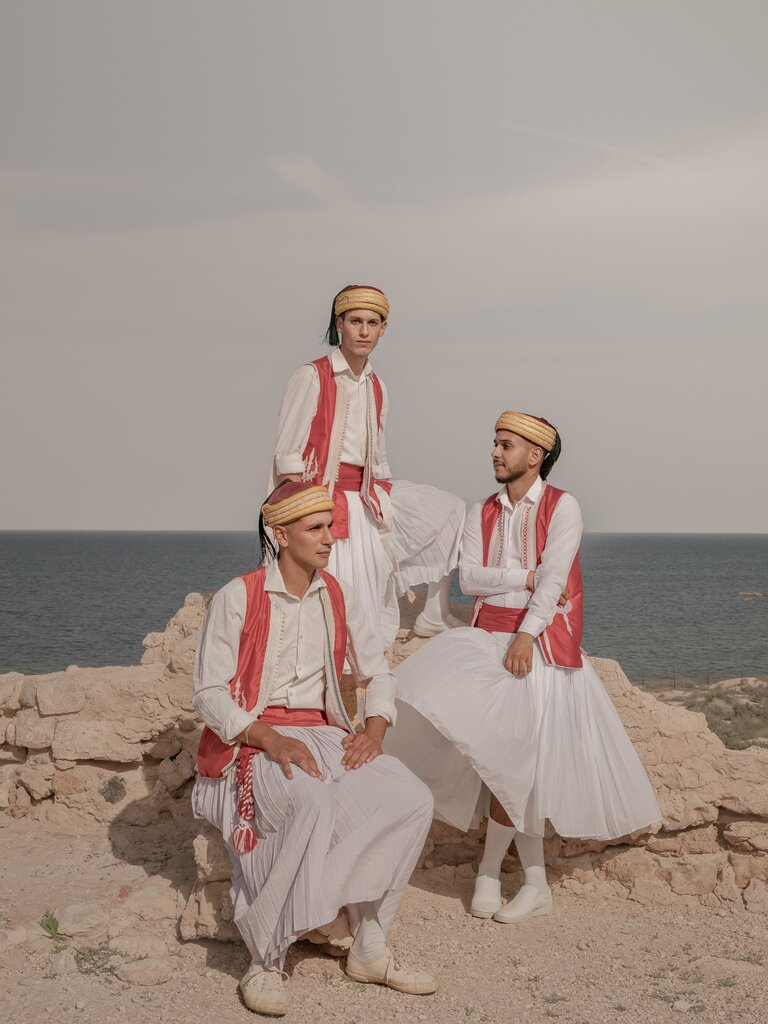 Three men in traditional dress pose by the ocean.