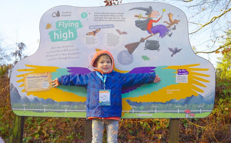 A young boy with arm spread as wings, in front of a panel in the forest.